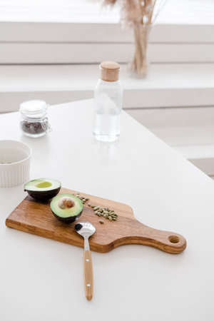 A still life sliced avocado with pumpkin seeds lies on a wooden board on a white table, against the...