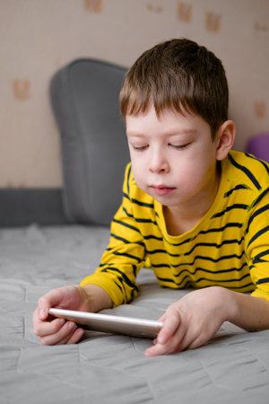 a teenage boy lies on the couch with a phone in his hands. The child plays at home in the phone. Recreation with the phone. Holidays with gadgets.の写真素材
