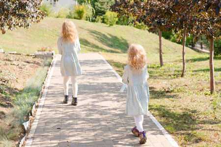 2 little blonde girls with curly hair are walking in a summer park. Cute and little girls on the street. 2 sisters walking outside in matching dresses and white tightsの写真素材
