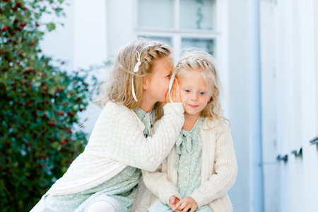 Two little cute girls are sitting in the garden and sharing secrets. One girl whispers something in another's ear. The two sisters have a close relationship.の写真素材