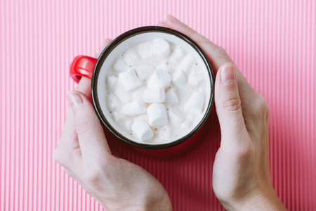 The girl's hands hold a large cup of cocoa or hot chocolate with marshmallows on a pink background, top viewの写真素材