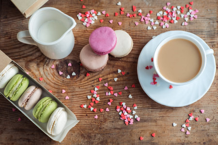 Macaroons in a box, a cup of coffee, a milk jug against the background of small hearts on a wooden background, top viewの写真素材