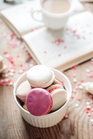Macaroons in a bowl and a cup of coffee, a book on the background of small hearts on a wooden...
