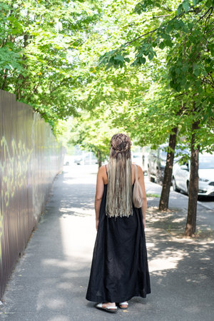 A girl in a black dress and long afro-braids walks along a summer street, rear viewの写真素材