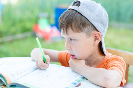 a boy in a cap solves math examples at a table on the street. Child doing homework in the parkの写真素材