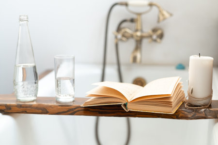 a book, a bottle of water, a glass and a candle on a wooden tray above the bathroom, against the background of an antique faucetの写真素材