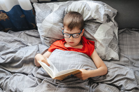 boy with glasses lies on a bed under a blanket and reads a book, top view. A child of primary school age is reading a book in his bed.の写真素材