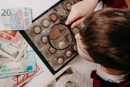 collecting coins The boy examines an old coin through a magnifying glass. Dollars, euros on the table. Money of different nations. Savings in the form of currencies and coinsの写真素材