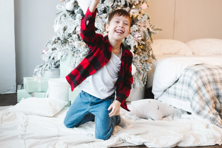 A boy in a red checkered shirt sits by the Christmas tree with gifts on the floor and fights with a pillowの写真素材
