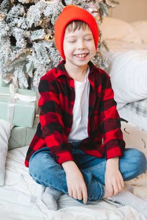 A boy in a red cap and a plaid shirt sits near a Christmas tree with gifts on the floor and smilesの写真素材