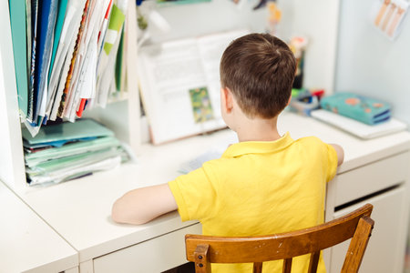 A boy in a yellow T-shirt sits and writes in a notebook at a table at home, rear view. Schoolboy...