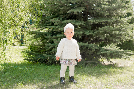 Cute blonde girl in a dress in pastel colors and white tights against the backdrop of the park. 1 year old baby learns to walkの写真素材