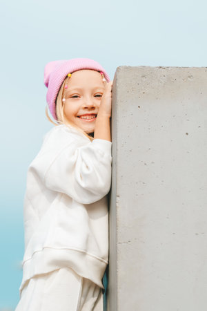 beautiful blonde girl in a white tracksuit and a pink hat climbs on a concrete fence, against the backdrop of a city landscapeの写真素材