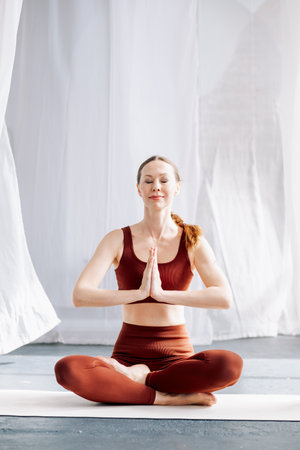 A slender woman in a brown sports uniform sits in the lotus position, with her eyes closed, against the background of white curtains and meditates. Yoga classes in the loft studioの写真素材