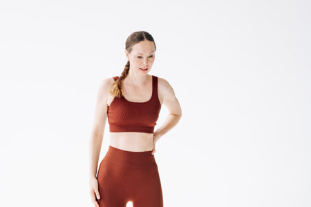 A slender woman in a brown sports uniform with a sore lower back holds her back, on a white...