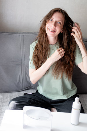 beautiful young woman with long wavy hair doing styling and combing it with a comb, looking in the mirror, sitting on the sofa, at homeの写真素材