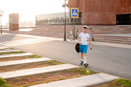 A 12-year-old boy in shorts and a T-shirt crosses the road next to a pedestrian crossing at sunset. A lonely teenager walks through the city in summer against the backdrop of houses.の写真素材