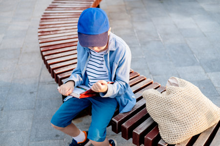a 10-year-old boy wearing glasses and a cap sits on a bench on the street and reads an interesting book against the sky, copy spaceの写真素材