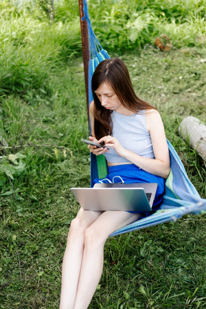 young woman with long hair and shorts sits in a hammock in the summer and works online on a laptop on the street. Freelancer on vacation, with a phone and laptop in his handsの写真素材