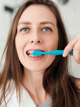 portrait of blue-eyed young woman with braces brushing her teeth with blue toothbrush.の写真素材