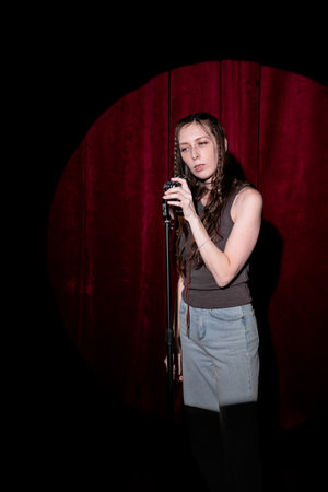 Young woman in jeans and a T-shirt standing behind a microphone on stage and singing. Performing a monologue on stageの写真素材