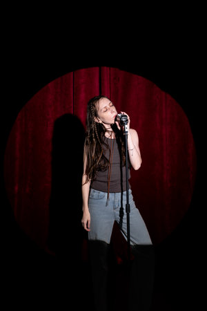 Young woman in jeans and a T-shirt standing behind a microphone on stage and singing. Performing a monologue on stageの写真素材