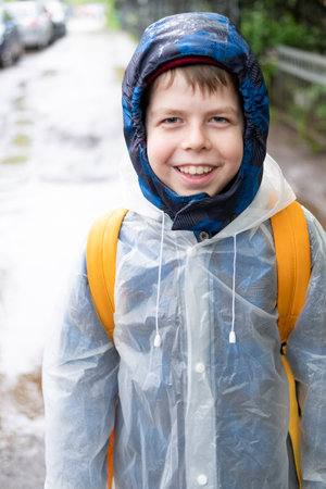 Portrait of smiling Boy 11 years old in transparent raincoat with yellow backpack standing outside during rain. Happy child on rainy dayの写真素材
