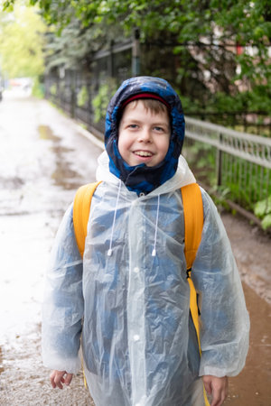 Portrait of smiling Boy 11 years old in transparent raincoat with yellow backpack standing outside during rain. Happy child on rainy dayの写真素材