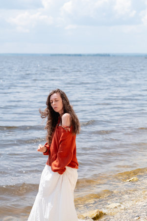 A young woman with long hair and blue eyes, in a white skirt and a terracotta sweater, stands barefoot in the water against the backdrop of the seaの写真素材
