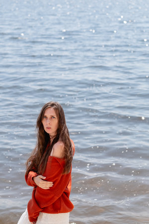A young woman with long hair and blue eyes, in a white skirt and a terracotta sweater, stands barefoot in the water against the backdrop of the seaの写真素材