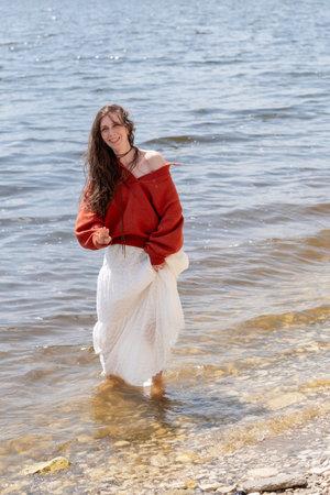 A young woman with long hair and blue eyes, in a white skirt and a terracotta sweater, runs barefoot on the water against the backdrop of the sea and laughsの写真素材