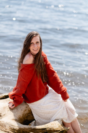 A young woman with braces, long hair and blue eyes, in a white skirt and a terracotta sweater, sits on a tree stump against the backdrop of the sea and smiles.の写真素材