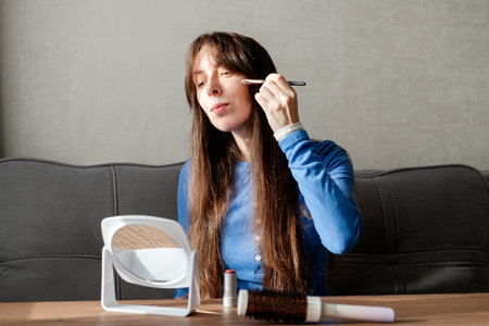 A young woman with long, damp hair applies eye makeup with a brush in front of a mirror while sitting on a sofa at a table. Morning routine before leaving the houseの写真素材