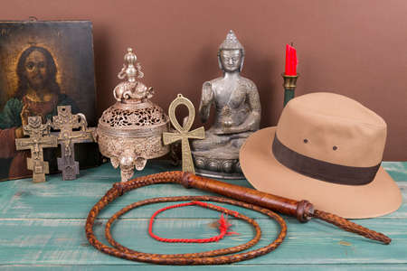 Adventure and archeological concept for lost artifacts with hat, whip, ancient iron vase, holy image, key of life, vintage crosses on green wood table and brown background.の写真素材