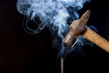 Abstract photo of metal rusty hammer above nail with smoke on black background.
