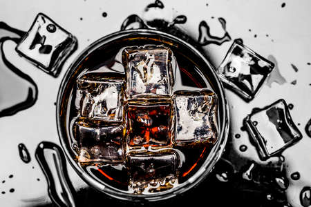 Glass with cola and ice cubes surrounded by ice cubes and water drops.