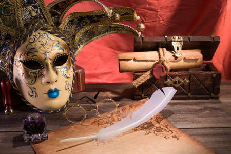 Theater concept. Vintage still life with quill and scroll near mask, old chest and glasses on red background.の写真素材