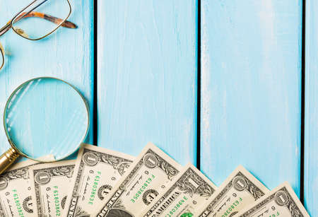 Magnifying glass with glasses and dollar bills on blue wooden background. Free space for text.