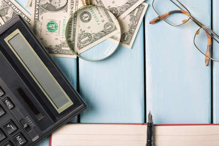 Business concept. Magnifying glass with glasses, dollar bills, notebook, fountain pen and calculator on blue wooden background.の写真素材