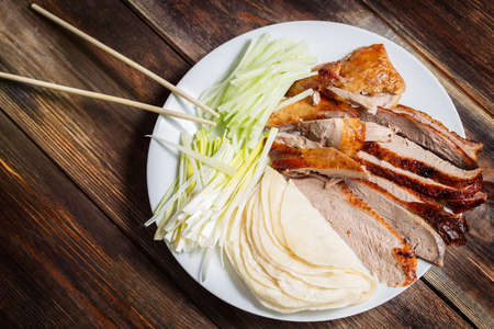 Peking Duck - Chinese roast duck with cucumber pieces, onion, pancakes on white plate close up. Wooden table and top view.の写真素材