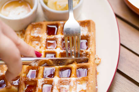 Woman hands cutting belgium waffles with fork and knife on plate with butter, maple sauce on wooden tableの写真素材