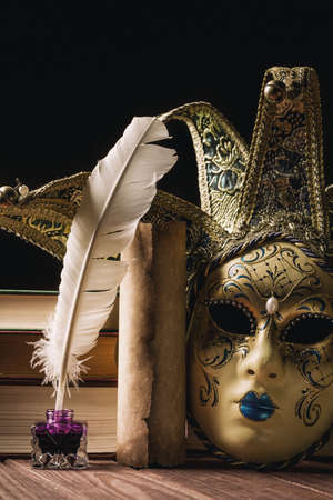 Literature concept. Inkstand with feather near venetian mask, books and old scroll on black background.の写真素材