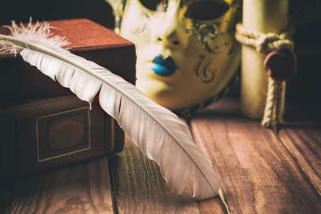 Literature concept. Feather on book near venetian mask and old scroll on wooden background.の写真素材