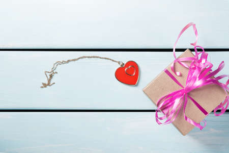 Gift box with nice pink ribbon and decorative heart with chain on blue wooden background. Free space for text.の写真素材