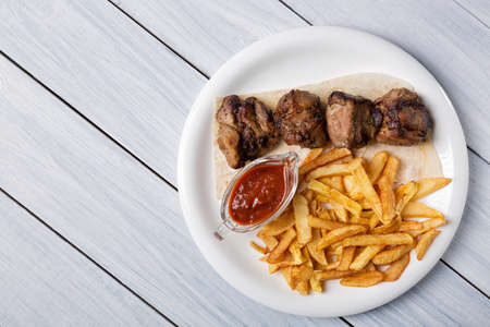 Grilled shish kebab served with french fries and sauce on white wooden table.