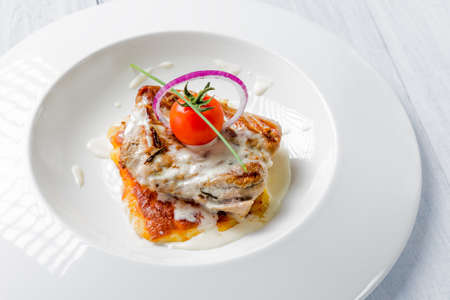 Tasty roasted pork pieces with cheese sauce, tomato and onion on white plate close up. Selective focus image.の写真素材