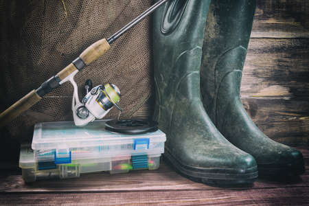 Fishing concept. Fishing tackles in boxes, rod, reel, fishing cage with rubber boots on timber board background. Toned image.の写真素材