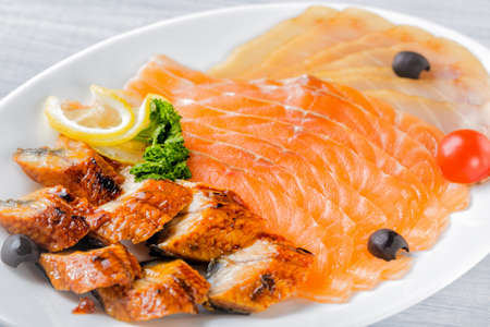 Salmon fillet pieces, conger eel, sturgeon pieces served with lemon, black olives, herbs and cherry tomato on white plate close up with selective focus.の写真素材