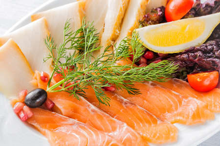 Salmon fillet pieces, sturgeon pieces served with lemon, black olives, herbs, cherry tomato and pomegranate seeds on white plate close up with selective focus.の写真素材