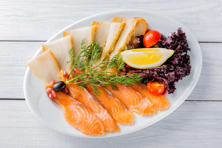 Salmon fillet pieces, sturgeon pieces served with lemon, black olives, herbs, cherry tomato and pomegranate seeds on white plate and wooden table. Top view.の写真素材